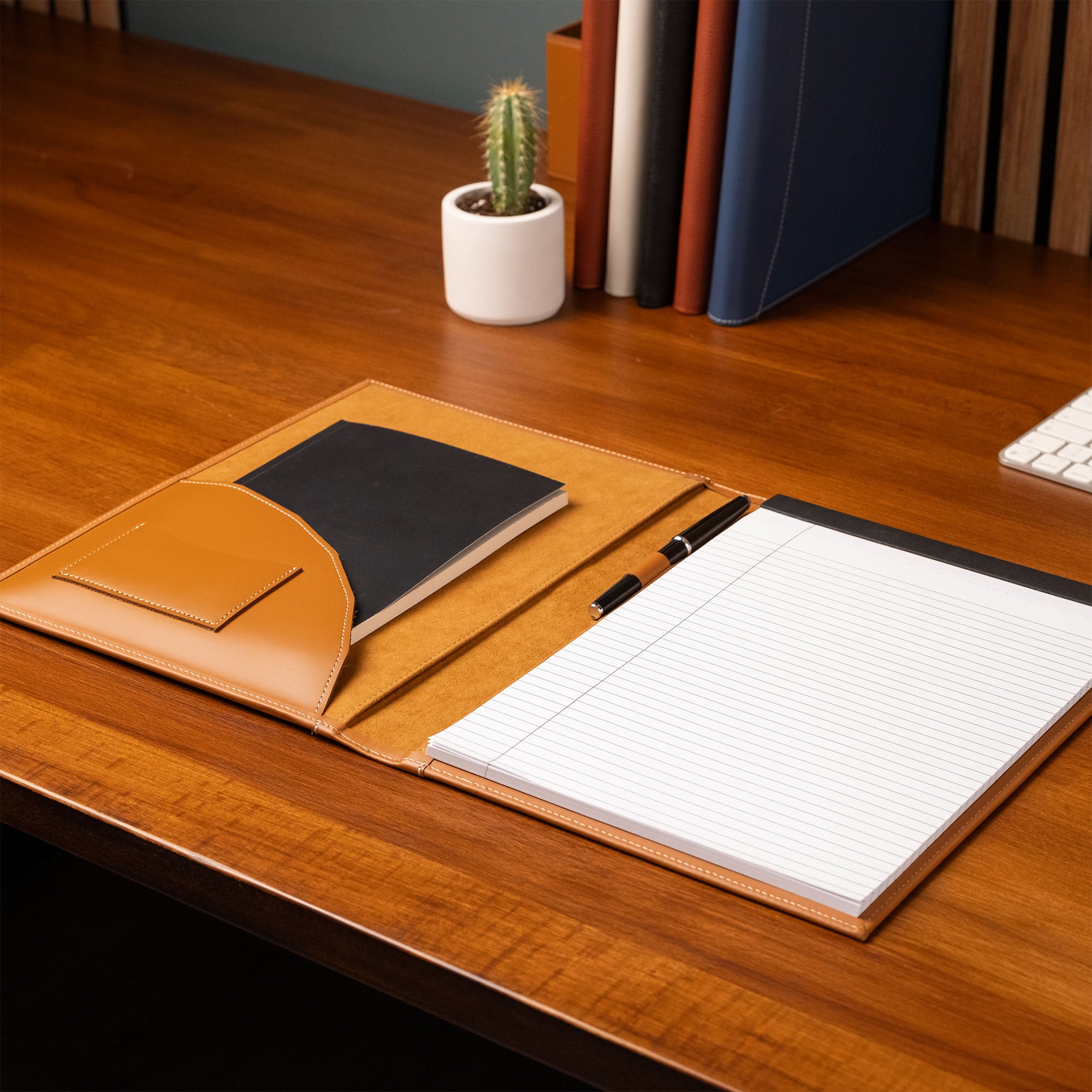 Notebook with pen on a wooden desk with books and a cactus in the
background